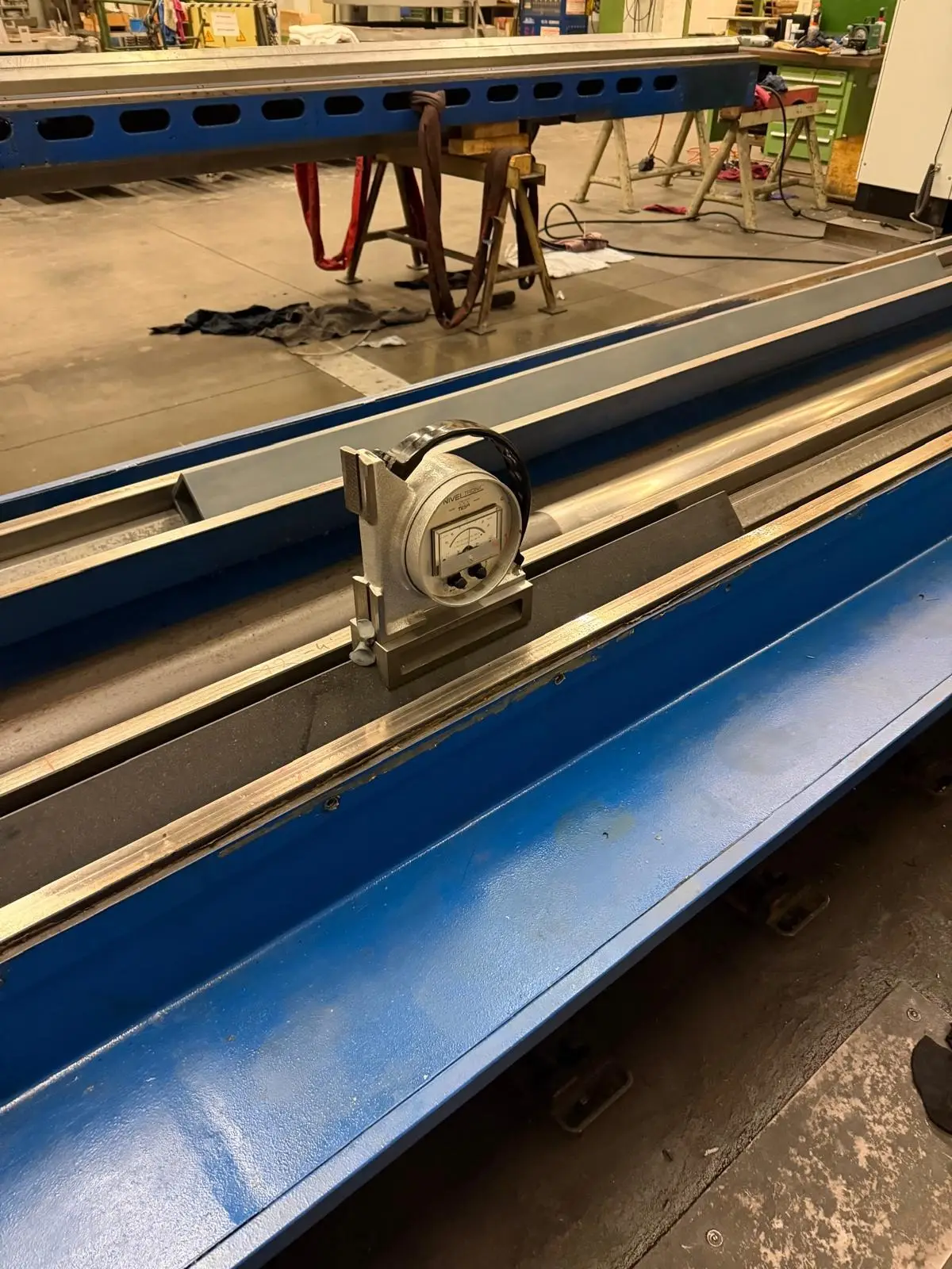 Elektronische Libelle NIVELTRONIC von TESA in ihrer Aufnahme auf einem dunklen Lineal, das in der Führung eines langen, blau lackierten Maschinenbetts liegt. Dahinter die Halle mit Böcken und Hebebändern.